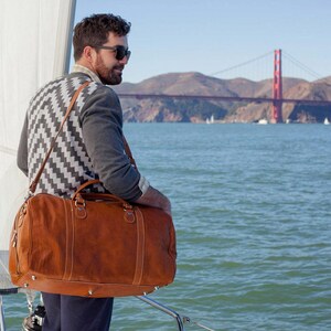 Pode incluir: Uma mala de viagem de couro castanho com uma al&ccedil;a de ombro. A mala tem um fecho de correr e tachas de metal na parte inferior. O fundo mostra uma ponte sobre a &aacute;gua.