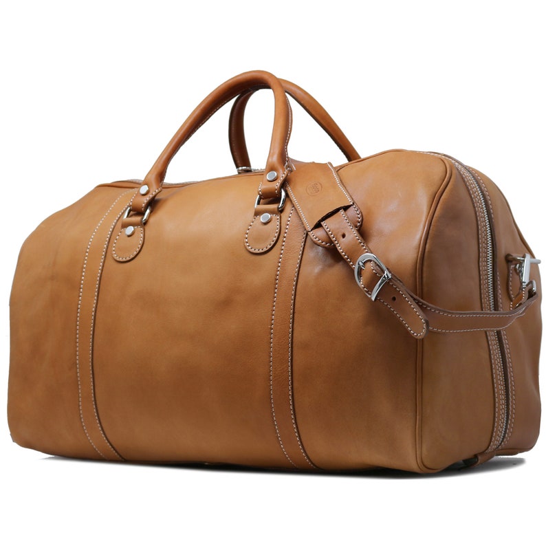 Puede incluir: Bolso de viaje de cuero marr&oacute;n claro con cierre de cremallera superior, dos asas superiores y una correa para el hombro desmontable. El bolso presenta costuras blancas y herrajes plateados. Dise&ntilde;ado para viajes, tiene un aspecto cl&aacute;sico y atemporal.