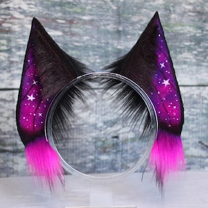 Puede incluir: Un par de orejas de gato de piel sintética negra y morada sobre una diadema transparente. Las orejas están pintadas con un diseño de galaxia, con estrellas blancas y un degradado de morado y rosa. Las puntas de las orejas son de color rosa brillante.