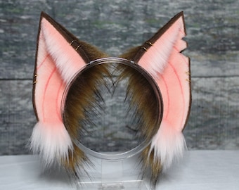 May include: A pair of brown and pink cat ears with white fur trim. The ears are attached to a headband and have gold wire accents.