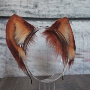 May include: A pair of brown and white faux fur ears attached to a headband. The headband is clear plastic and has a metal band.