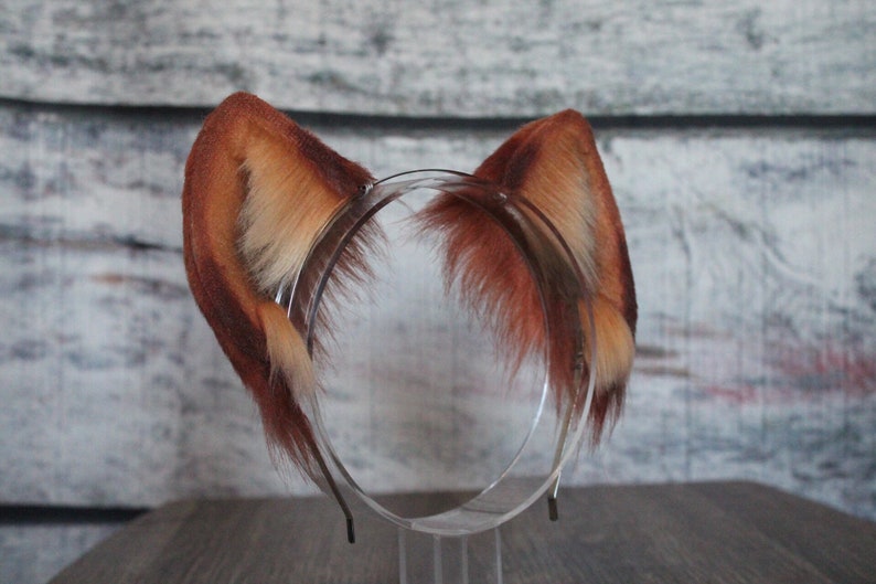 May include: A pair of brown and white faux fur ears attached to a headband. The headband is clear plastic and has a metal band.