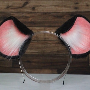 May include: A pair of cat ears on a clear headband. The ears are black on the outside, with pink and white fur on the inside. The headband is made of clear plastic and metal.