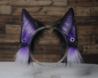 May include: A pair of purple and white cat ears with black fur and silver accents. The ears are attached to a headband.