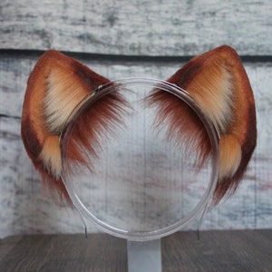 May include: A pair of brown and white faux fur cat ears headband. The headband is clear plastic and the ears are soft and fluffy.