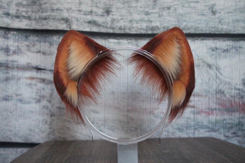 May include: A pair of brown and white faux fur cat ears headband. The headband is clear plastic and the ears are soft and fluffy.