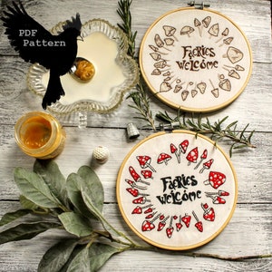Könnte beinhalten: Zwei Stickrahmen mit weißem Stoff und braunen Holzrahmen. Der Stoff in jedem Rahmen ist mit einem Stickmuster aus Pilzen mit roten Hüten und weißen Punkten bestickt. Der Text "Fairies Welcome" ist in braunem Garn in der Mitte jedes Rahmens gestickt.