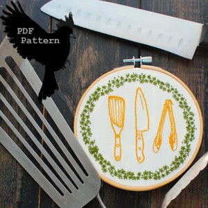 May include: Embroidered kitchen tools on a white fabric background in a wooden embroidery hoop. The tools include a spatula, a knife, and tongs. The hoop is surrounded by a green embroidered border.