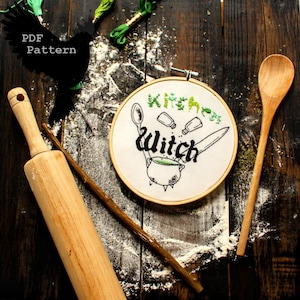 May include: A white embroidery hoop with the text "Kitchen Witch" embroidered in black thread. The design includes a cauldron, herbs, and a spoon. The hoop is on a wooden surface with flour scattered around it.
