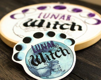 Puede incluir: Un aro de bordado blanco con las palabras "Lunar Witch" en escritura negra y un diseño de fases lunares púrpura. Una pegatina con el mismo texto y un diseño de fases lunares acuarela azul y blanco está sobre una superficie de madera.