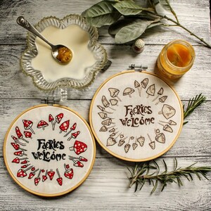 May include: Two embroidery hoops with a white background and brown stitching. One hoop features a circle of red mushrooms with white spots and the text "Fairies Welcome". The other hoop features a circle of brown mushrooms with the text "Fairies Welcome".