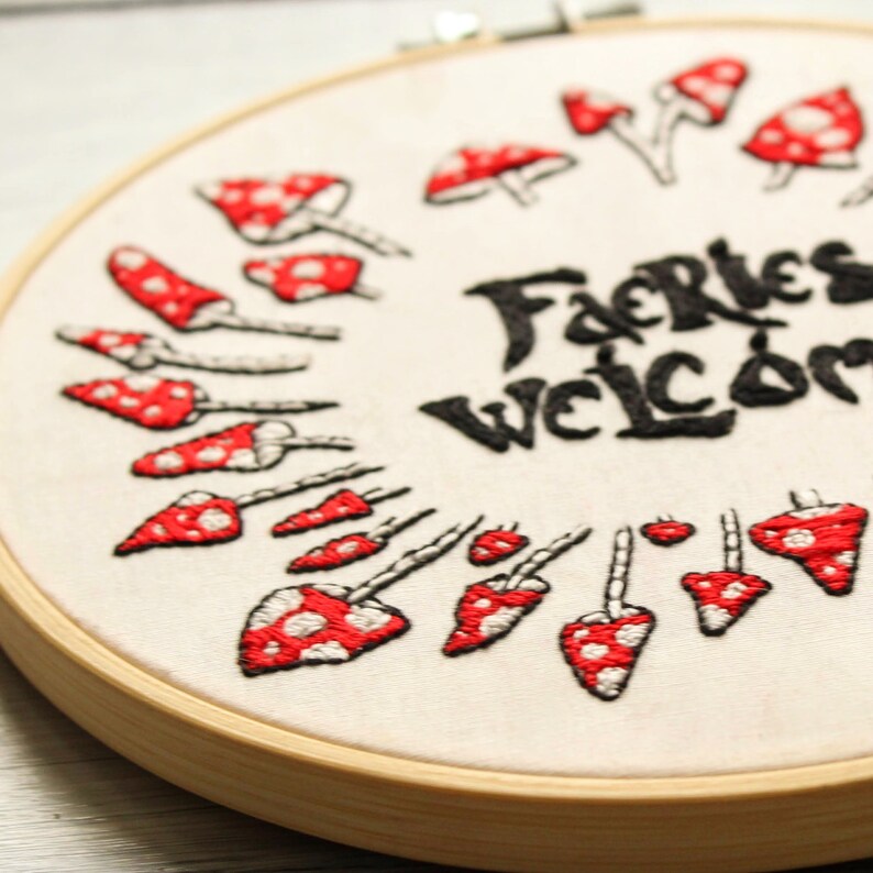 May include: Embroidered hoop art featuring the text "Fairies Welcome" in black cursive lettering, surrounded by a circle of red and white mushroom illustrations.
