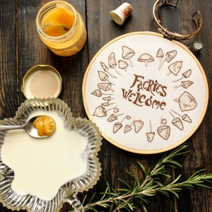 May include: A white embroidery hoop with brown thread embroidery of mushrooms and the text "Fairies Welcome". The hoop is on a wooden surface with a jar of honey, a small glass jar, and a sprig of rosemary.