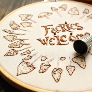 May include: Embroidered hoop art featuring the text "Fairies Welcome" in brown thread. The hoop is encircled by a ring of brown and white embroidered mushrooms.