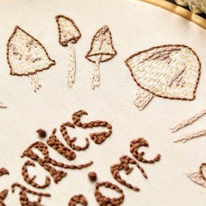 May include: Embroidered hoop art featuring a white background with brown and beige mushrooms and the text "Shrooms for the soul".