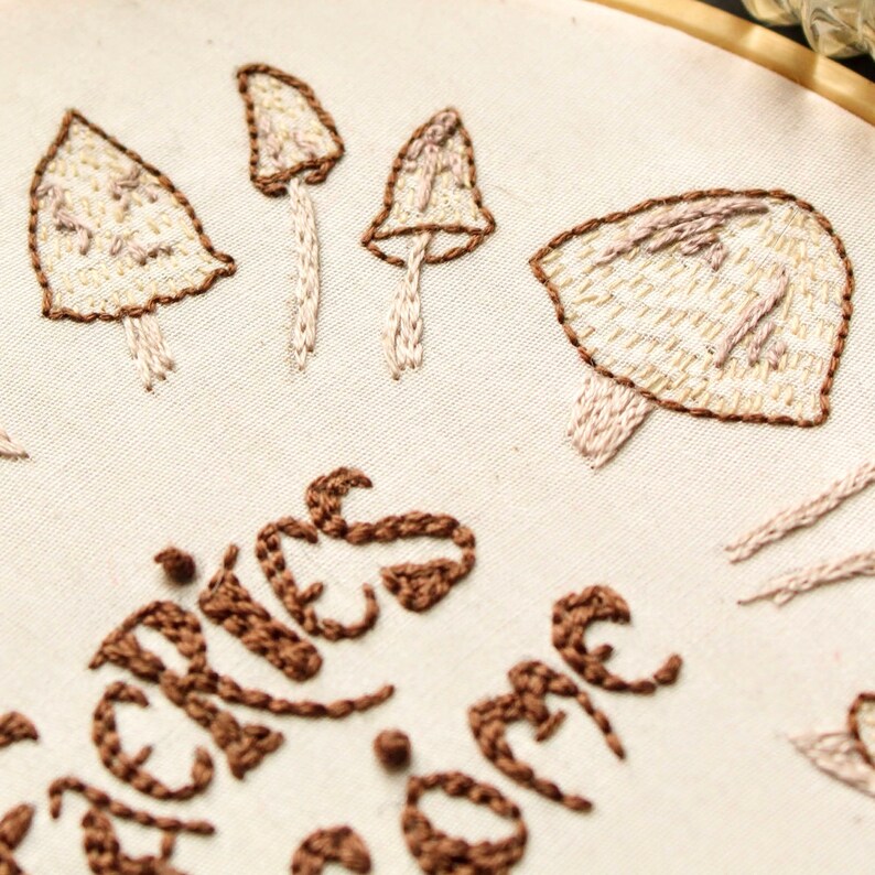 May include: Embroidered hoop art featuring a white background with brown and beige mushrooms and the text "Shrooms for the soul".