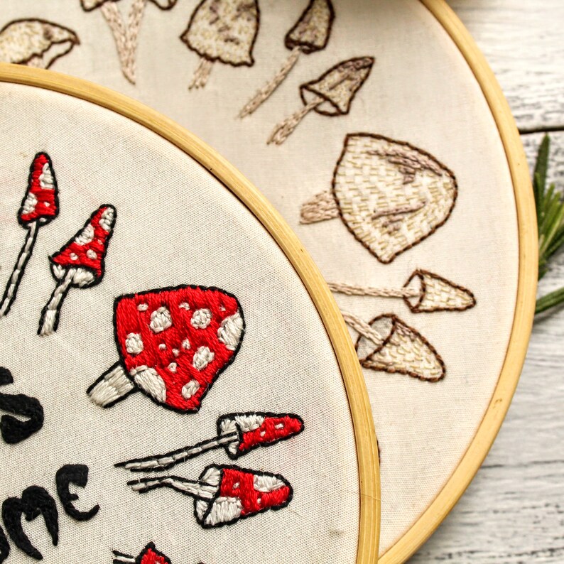 May include: Two embroidery hoops with different mushroom designs. One hoop features red and white mushrooms with white polka dots and the text "Shroom Queens". The other hoop features beige mushrooms with brown stems.