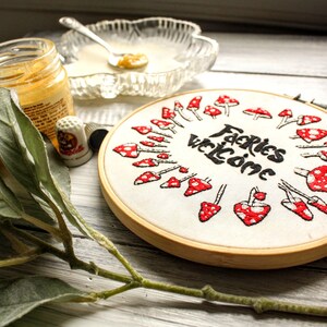May include: A white embroidery hoop with a red and white mushroom design and the text "Fairies Welcome" in black lettering.