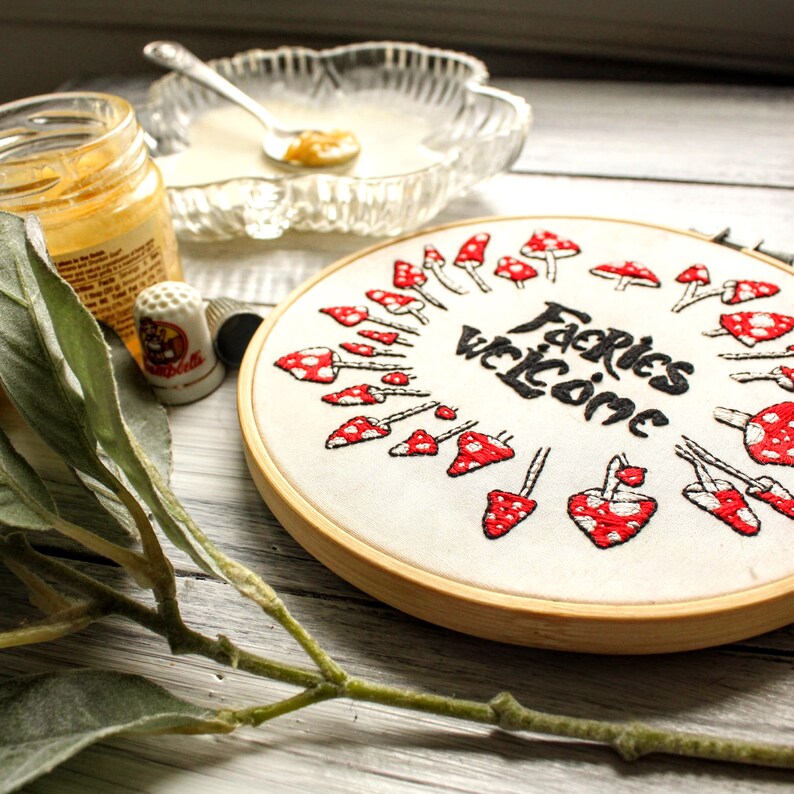 May include: A white embroidery hoop with a red and white mushroom design and the text "Fairies Welcome" in black lettering.