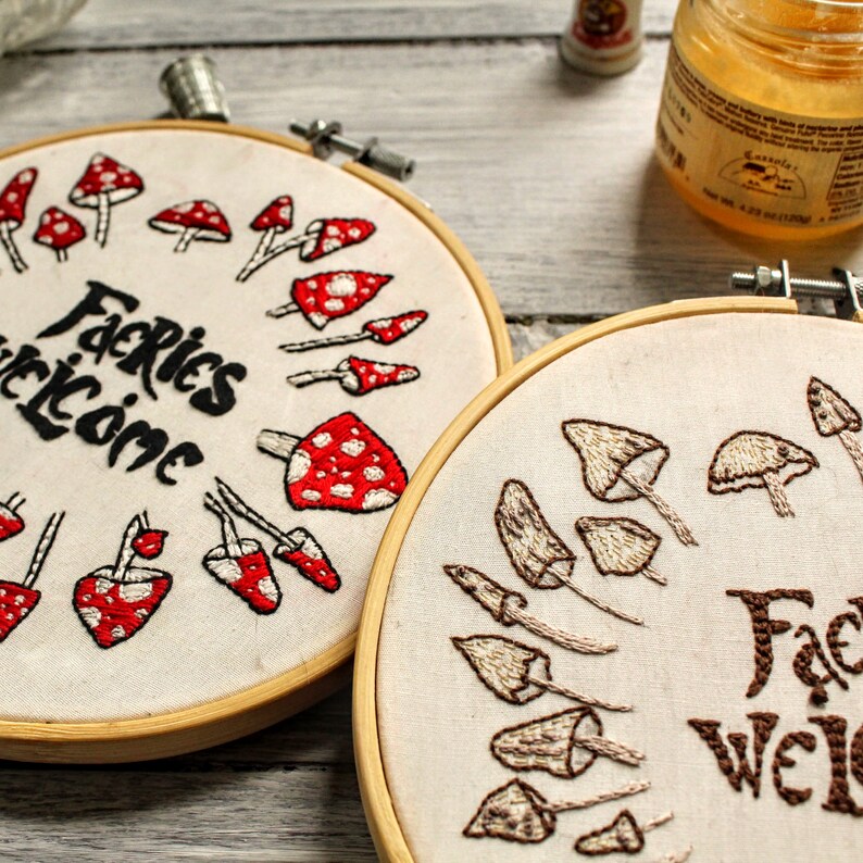 May include: Two embroidery hoops with white fabric and wooden frames. One hoop features red and white mushrooms with the text "Fairies Welcome". The other hoop features brown mushrooms with the text "Fairies Welcome".