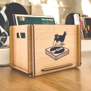 May include: A wooden record storage box with a black illustration of a bird on a record player. The box holds a record album with the text "Blues Brothers". The box is made of light-colored wood and has a handle.