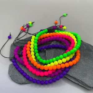 May include: A set of colourful bracelets made with neon beads and black string. The bracelets are arranged in a rainbow pattern, with purple, pink, orange, yellow, green, and blue beads.