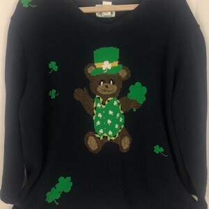 May include: A navy blue sweater with a brown bear wearing a green hat and holding a shamrock. The sweater is decorated with green shamrocks.