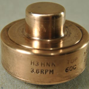 Può includere: Componente meccanico color rame con corpo cilindrico e cilindro più piccolo rialzato sulla parte superiore. Il testo "H3 HNK 3.6RPM TOP 60C" è inciso sul lato. L'oggetto ha una lucentezza metallica e poggia su una superficie neutra.