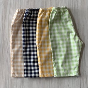 May include: Four pairs of elastic-waist shorts featuring a gingham check pattern in beige, navy blue, yellow, and light green. Each pair of shorts is displayed side-by-side, showcasing the consistent checkered design and the gathered waistband.