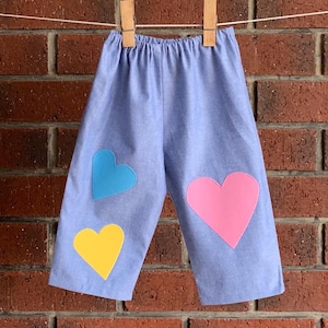May include: Light blue trousers featuring an elastic waistband and three heart-shaped fabric appliqués. The left leg displays a small blue heart above a yellow heart, while the right leg features a single large pink heart centred on the fabric.
