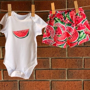 May include: A white baby onesie with a watermelon graphic and a pair of red and green watermelon print shorts.