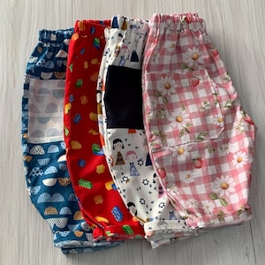 May include: Four pairs of elastic-waist trousers featuring distinct patterns: blue with white semicircles, red with colourful building blocks, white with illustrated figures and a black patch, and pink gingham with white daisies and red strawberries.
