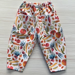 May include: White trousers featuring an all-over colourful print of various vegetables including tomatoes, carrots, radishes, and sliced limes. The trousers have an elasticated waistband and two patch pockets on the back, showcasing a vibrant garden-themed pattern.