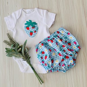 May include: A white baby bodysuit with a strawberry applique and matching blue gingham shorts with strawberry and daisy print. The outfit is designed for infants and toddlers.