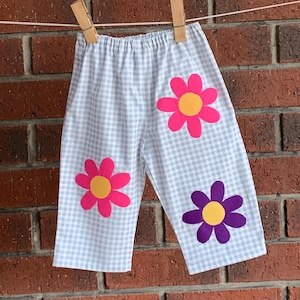 May include: Light grey and white gingham trousers featuring an elasticated waistband and three large flower appliqués. Two flowers are bright pink with yellow centres, and one flower is purple with a yellow centre, all stitched onto the fabric.
