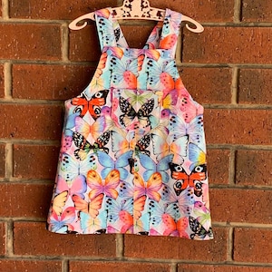 Puede incluir: Un vestido de peto blanco con estampado de mariposas de colores. El vestido tiene tirantes ajustables y un solo bolsillo en la parte delantera.