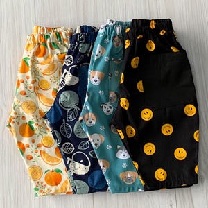 May include: Five pairs of patterned shorts are shown. The patterns include oranges, abstract shapes, dog faces, and yellow smiley faces on a black background. The shorts are made of a lightweight fabric.