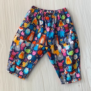 May include: Pull-on trousers with an elastic waistband featuring a dark blue background covered in a colourful, repeating pattern of various cartoon animals including rabbits, bears, and hedgehogs in shades of orange, pink, yellow, and light blue.