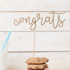 May include: A stack of chocolate chip cookies with a gold glitter "Congrats" topper on top. The cookies are on a white napkin and a glass of milk is in the background.