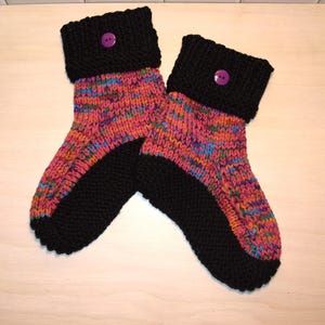 May include: A pair of hand-knitted slipper socks. The socks feature a black foot and cuff, with a colorful speckled pattern in the middle. Each cuff is adorned with a purple button. These cozy socks are perfect for lounging at home.