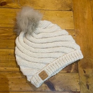 May include: A white knitted beanie with a fluffy pom pom on top. The beanie has a ribbed texture and a small brown tag on the side.