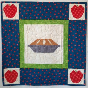 Peut inclure: Une tenture murale matelassée bleue et blanche avec un cadre vert. Le centre de la tenture présente une tarte brune et blanche avec une croûte grise. Les coins de la tenture sont décorés de pommes rouges à pois blancs.