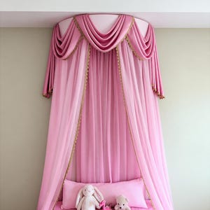 May include: A pink bed canopy featuring draped fabric with gold trim, suspended from a circular mount. Below the canopy, a pink pillow and pink bedding are arranged with a plush rabbit, a small pink dinosaur, and a small plush bear toy.