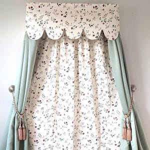 May include: A window treatment featuring a valance with a scalloped edge and a floral pattern of red flowers and green leaves on a cream background. The curtains are a light green color and are tied back with tassels.