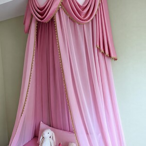 May include: A pink canopy bed curtain featuring a semi-circular top valance with draped fabric and gold lace trim. The sheer pink curtains hang vertically from the ceiling mount, creating a soft enclosure over a pink surface with three plush toys.