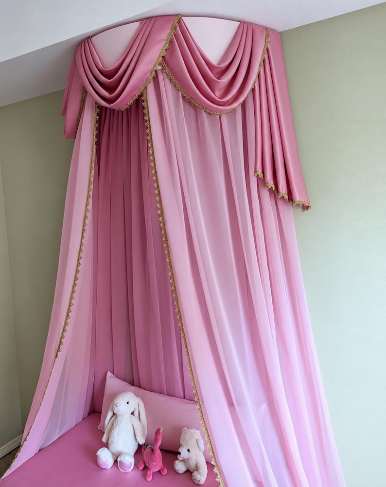 May include: A pink canopy bed curtain featuring a semi-circular top valance with draped fabric and gold lace trim. The sheer pink curtains hang vertically from the ceiling mount, creating a soft enclosure over a pink surface with three plush toys.