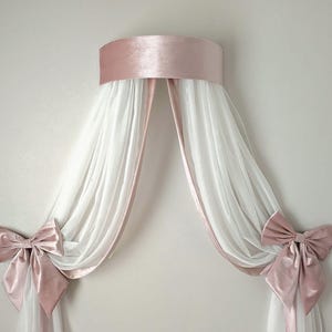Könnte beinhalten: Ein weißer Baldachin mit rosa Samtbesatz und Schleifen. Der Baldachin ist mit einem rosa Samtband an der Wand befestigt.