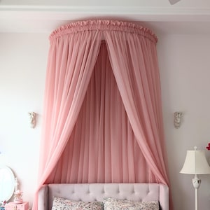 May include: A pink sheer canopy draped over a bed with a white headboard and floral patterned pillows. The canopy is gathered at the top and hangs down in soft folds. A pillow with the word "home" in gold lettering is on the bed.