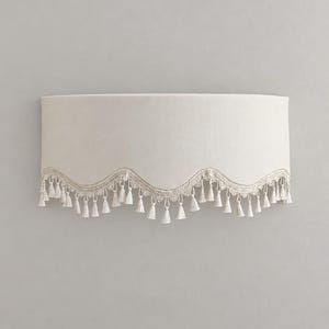 May include: A semi-circular off-white fabric lampshade featuring a scalloped bottom edge. The lower border is decorated with intricate lace trim and a series of small, hanging off-white tassels that follow the curved line of the shade.