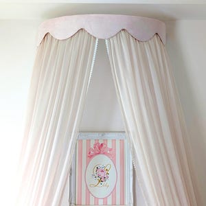 Könnte beinhalten: Ein weißer Baldachin mit einem rosa gewellten Rand hängt von der Decke. Ein weißer Rahmen mit einem rosa-weiß gestreiften Hintergrund und einem floralen Monogramm mit dem Namen "Lily" hängt an der Wand.